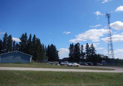 Yellow Quill First Nation is located in Rose Valley, Saskatchewan that spans across a total area of 571 square meters. The image contains a side shot of a blue building on the left with a parking lot to the right. There is a cell tower on the opposite side of the parking lot. There is some large tree cover on the side opposite the camera with a blue sky behind it.