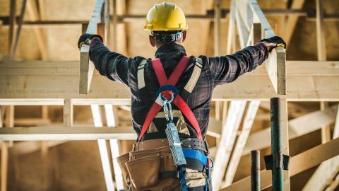 A construction worker is standing in a framed building with his back to the camera and a yellow hard hat on. 