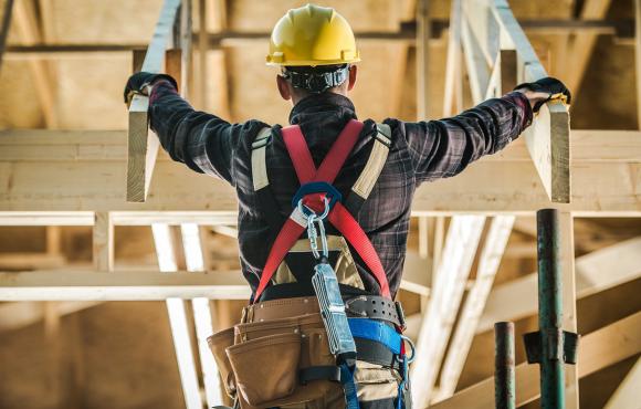 A construction worker is standing in a framed building with his back to the camera and a yellow hard hat on. 