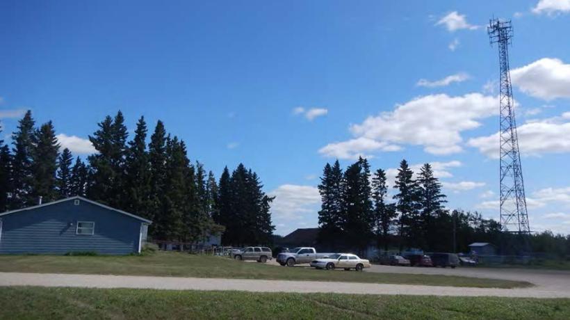 Yellow Quill First Nation is located in Rose Valley, Saskatchewan that spans across a total area of 571 square meters. The image contains a side shot of a blue building on the left with a parking lot to the right. There is a cell tower on the opposite side of the parking lot. There is some large tree cover on the side opposite the camera with a blue sky behind it.